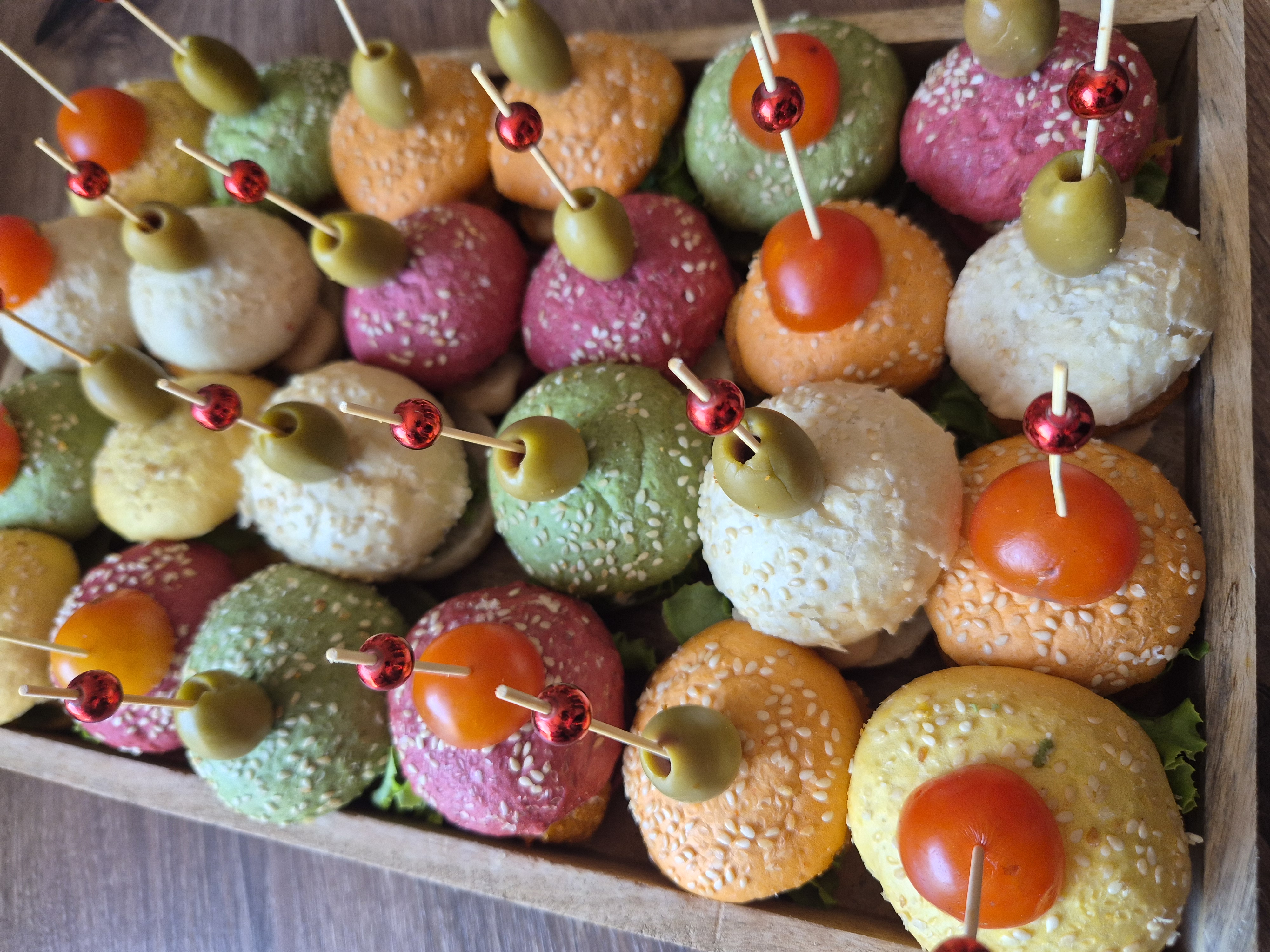 Barviti mini burgerji v roza, zeleni in oranžni barvi na lesenem pladnju – Prigrizki.si finger food